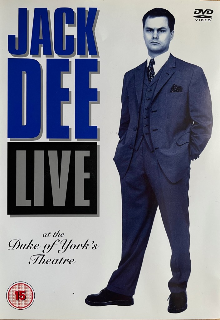Front cover of the DVD for Jack Dee Live at the Duke of York's Theatre, with a white background. The words Jack Dee Live are in very large letters on the left of the cover, his name in blue, while Live is in grey in a black box. The venue name is in much smaller curly handwriting style text. Alongside the title on the right is a full length blue-tinted photo of Jack wearing a dark coloured suit and tie with a white shirt. He is standing with his hands behind his back.