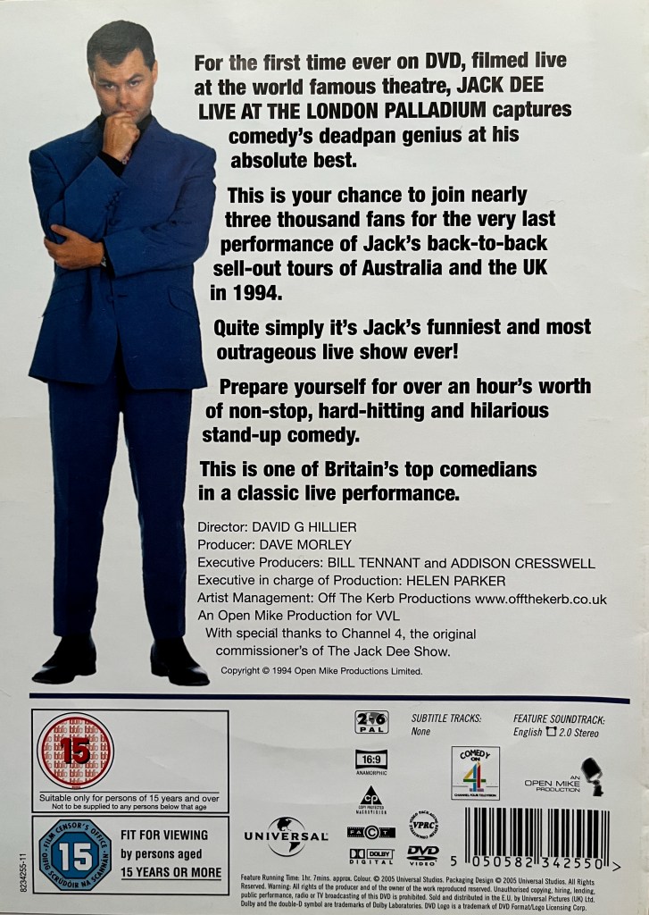 Back cover of the DVD for Jack Dee Live At The London Palladium, with a white background. It shows a full length photo of Jack on the left wearing a dark blue suit, his right hand resting thoughtfully on his chin while his left arm is folded across his chest to support his right elbow. A description and credits for the DVD are shown to the right of him, while technical and copyright information is across the cover below him at the bottom.