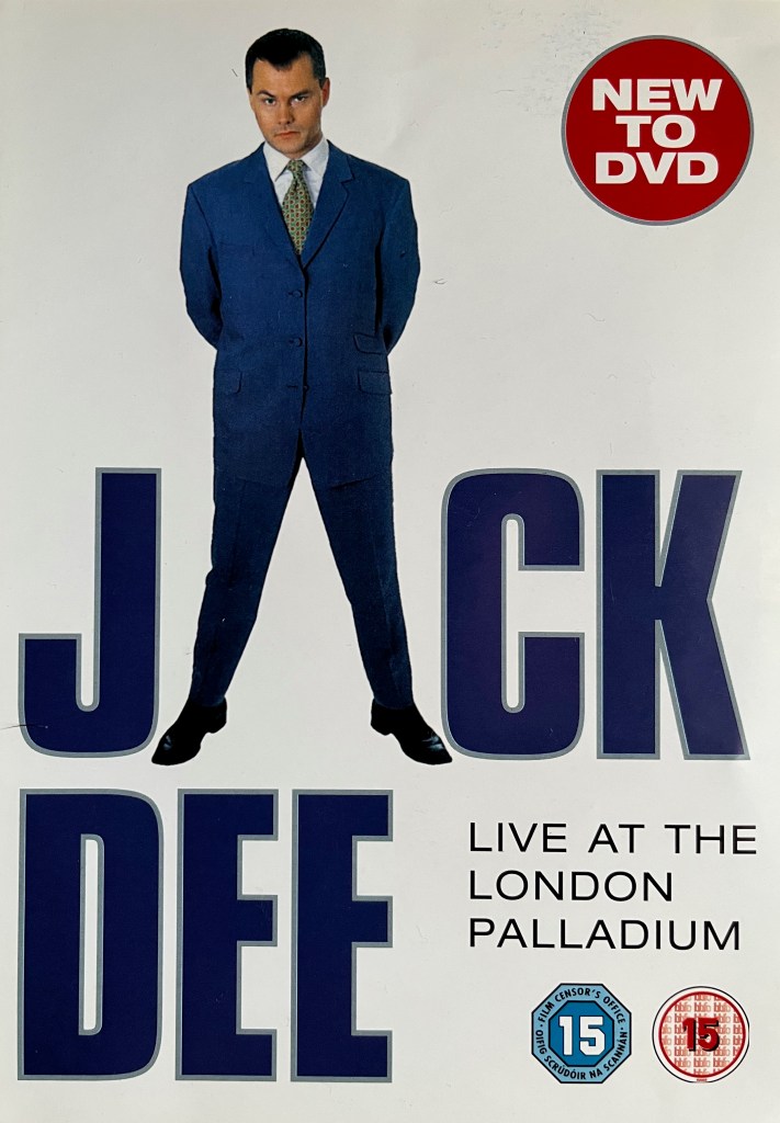 Front cover of the DVD for Jack Dee Live At The London Palladium, with a white background. It shows a full length photo of Jack in the top half, wearing a dark blue suit over a patterned tie and a white shirt. He is standing with his hands behind his back. His legs are apart as well, and they're used as a substitute for the letter A in the name Jack Dee beneath him, in large blue letters, forename on top and surname underneath. As his surname is shorter, the text Live At The London Palladium is in smaller black capitals in bottom right space on the cover.