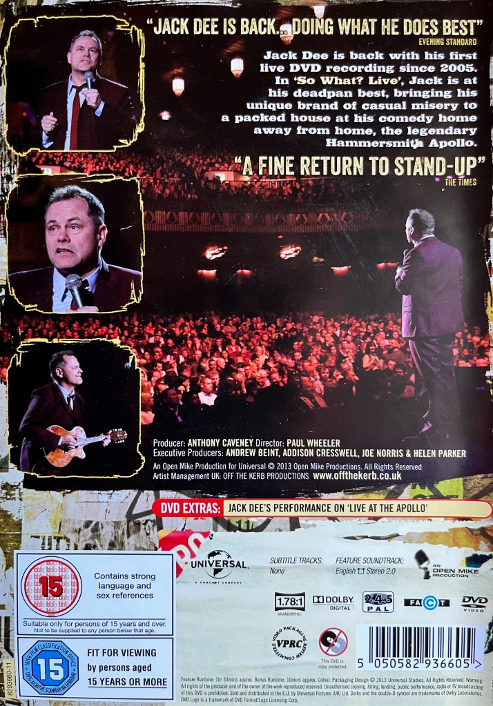 Back cover of the DVD for Jack Dee, So What Live. Beneath a short description at the top right, the photo dominating the cover is a view of Jack performing to the audience, as seen from the rear of the stage. On the left are 3 boxes showing a front view of Jack as he performs, two of which show him talking with a microphone, while the other shows him holding a guitar. The technical and copyright information is over a white background across the bottom.