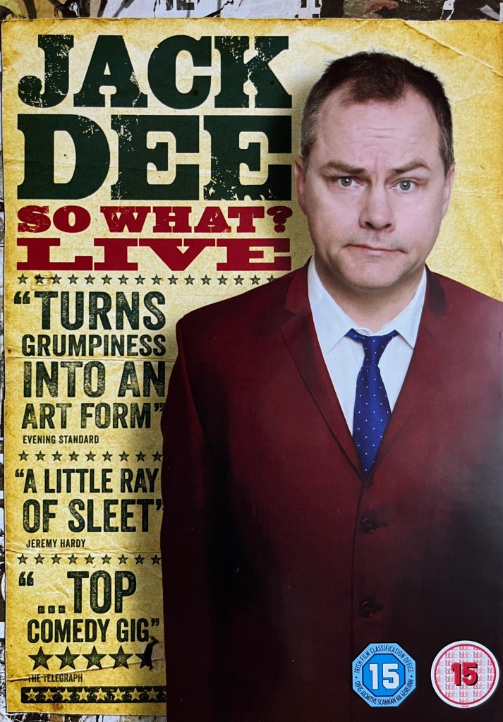 Front cover of the DVD for Jack Dee, So What Live. On the right is a half-length picture of Jack wearing a red jacket over a blue tie and a white shirt. At the top left is Jack's name in black and the show's title in red. Below that are a few review quotes in large black lettering. The Evening Standard says he turns grumpiness into an art form. Jeremy Hardy says he's a little ray of sleet. And the Telegraph, giving it 5 stars, says it's a top comedy gig.