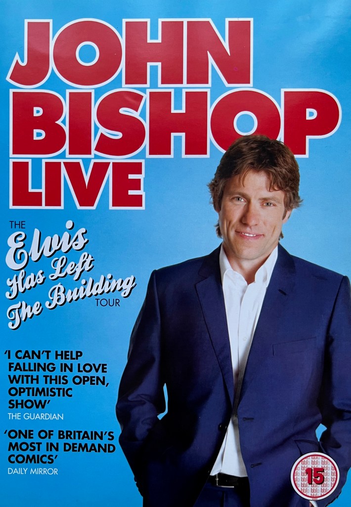 Front cover of John Bishop's DVD Elvis Has Left The Building. It shows John in front of a light blue background, smiling while wearing a dark blue jacket over a white shirt.