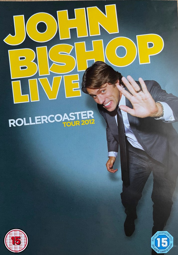 Front cover of John Bishop's DVD called Rollercoaster. It shows John wearing a grey suit and tie with a white shirt. He's hunched over slightly and is holding his hand up ahead of him as if signalling for something to stop.