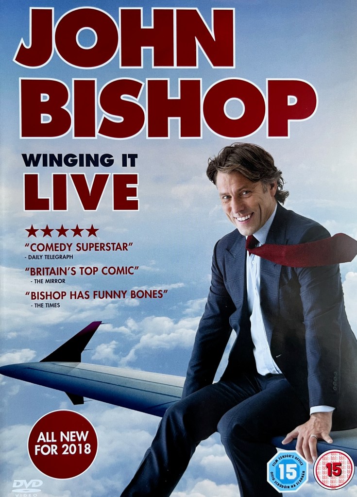 Front cover of John Bishop's DVD called Winging It. It shows John in a dark grey suit with a white shirt, smiling as he sits on the wing of a plane above clouds in the sky, his red tie being blown to the right by the wind.