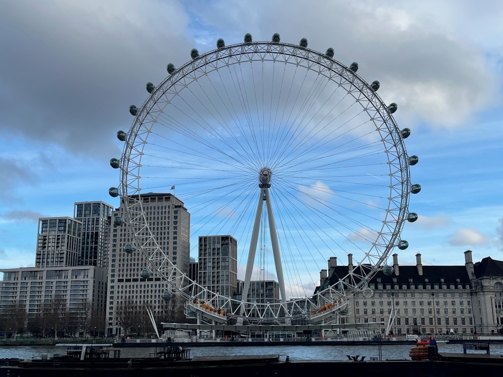 The huge London Eye observation wheel overlooking the River Thames, dwarfing the buildings behind it.