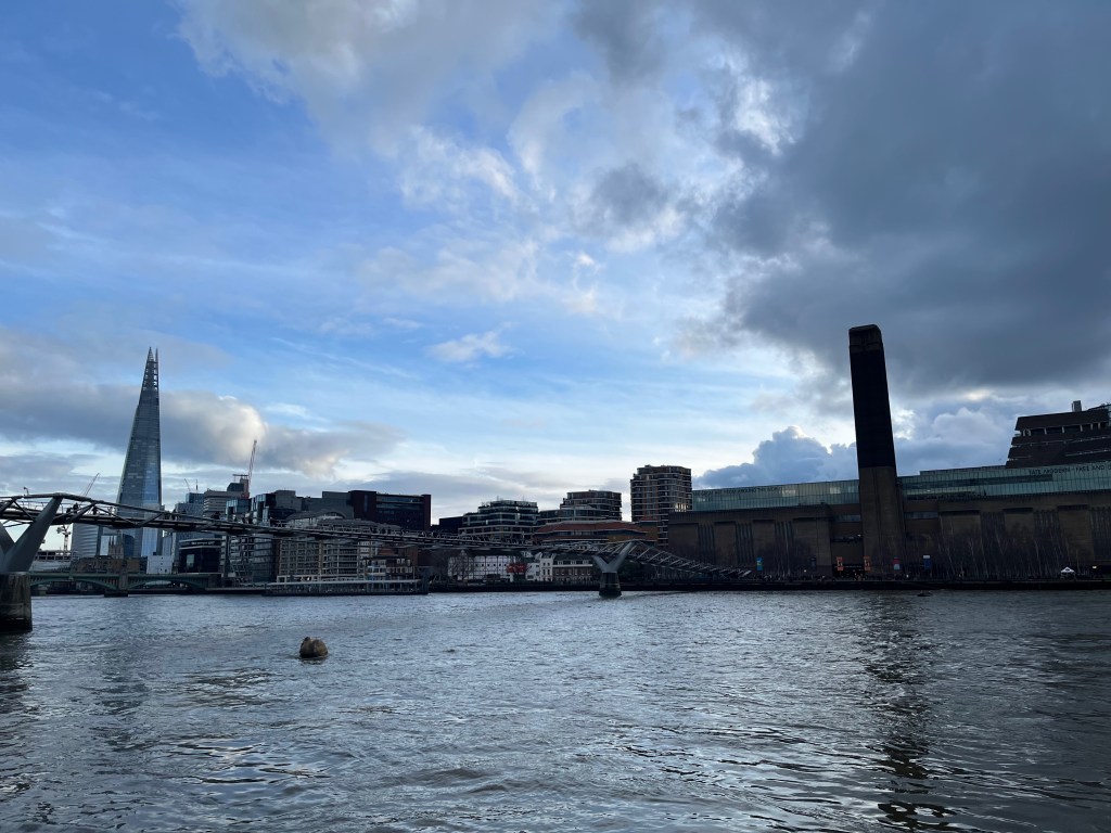 View across the River Thames to the south bank, with the tall pointed Shard building on the left, and the Tate Modern with its tall chimney on the right, while the Millennium Bridge crosses the water near to the latter.