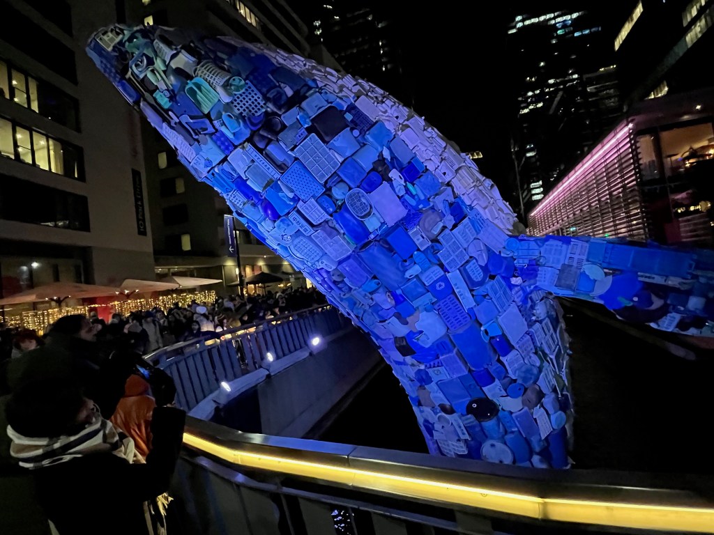 A huge sculpture of a blue whale lit up in blue, made out of plastic waste from the seas, sticking out of a dock in Canary Wharf, towering over the people walking on the path next to it.