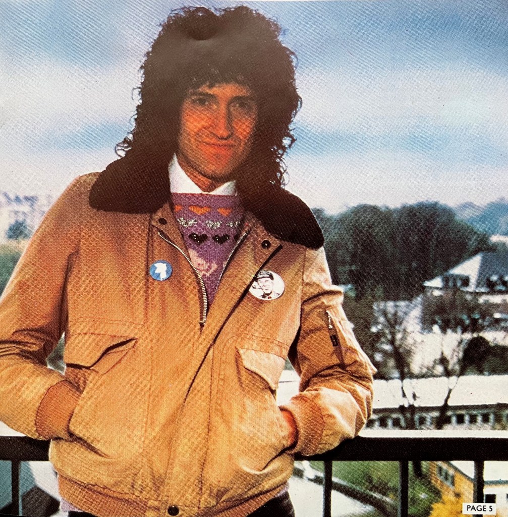Brian May, with his hands in his light brown jacket pockets, leaning back against a railing overlooking a snowy landscape with trees and a couple of buildings.