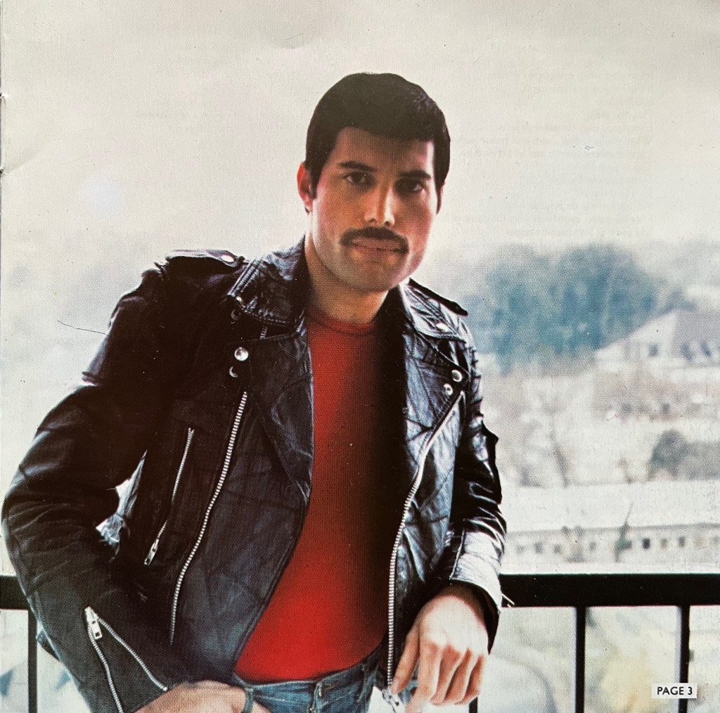 Freddie Mercury, sporting a moustache and wearing a black leather jacket over a red top, leaning back against a railing overlooking a snowy landscape with trees and a couple of buildings.