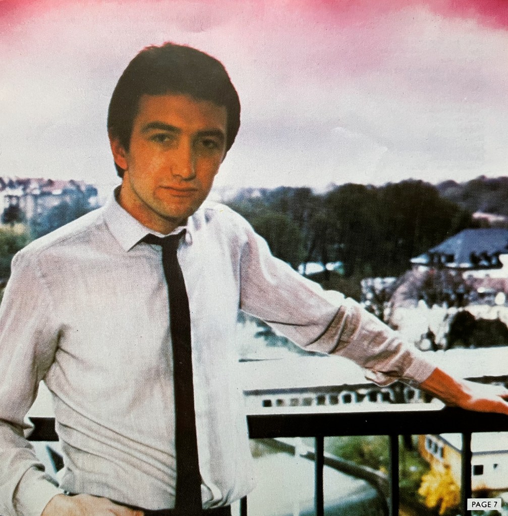 John Deacon, wearing a white shirt and a long black tie, leaning back against a railing overlooking a snowy landscape with trees and a couple of buildings.