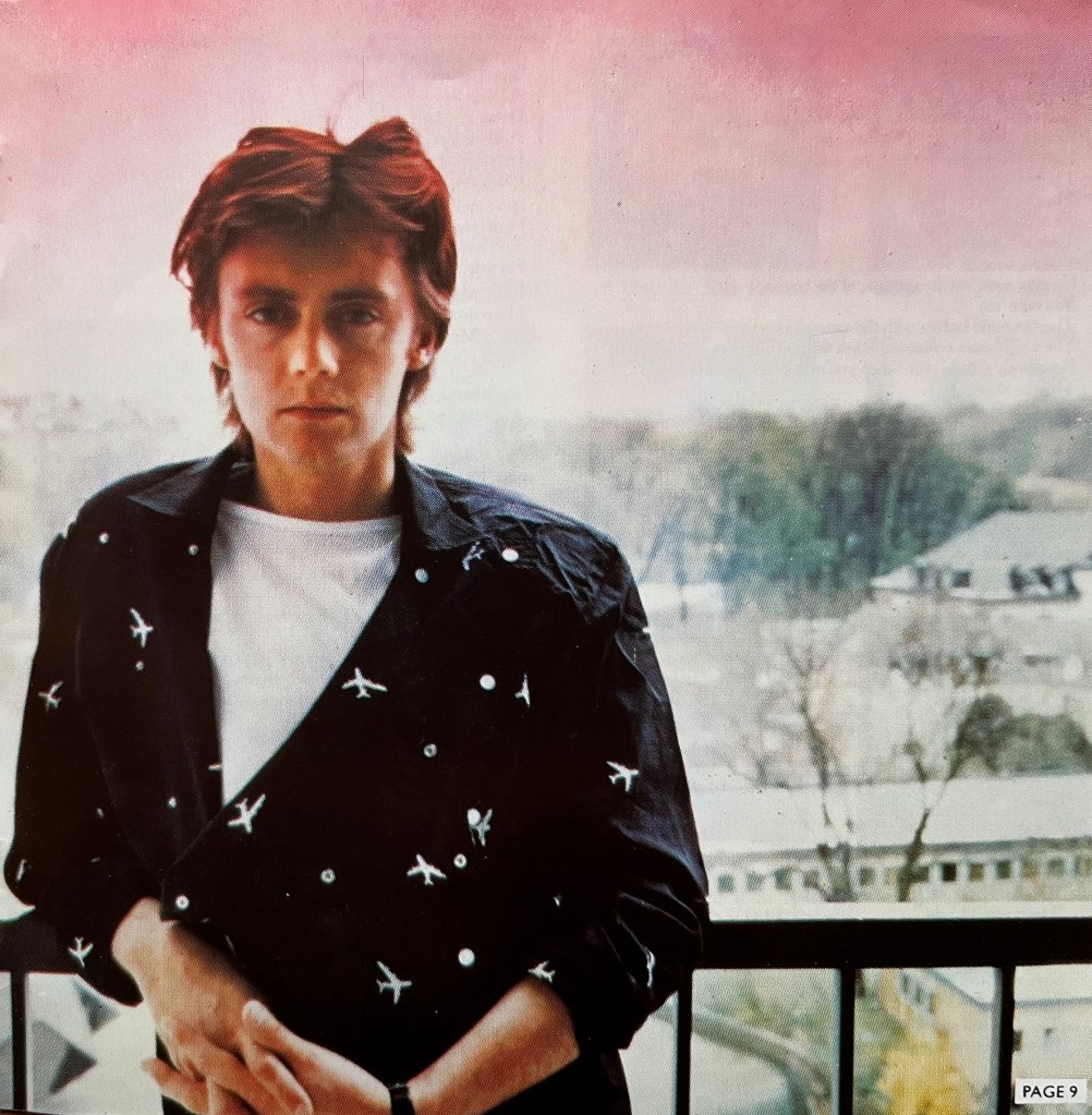 Roger Taylor, wearing a black jacket decorated with small white motifs of aeroplanes, leaning back against a railing overlooking a snowy landscape with trees and a couple of buildings.