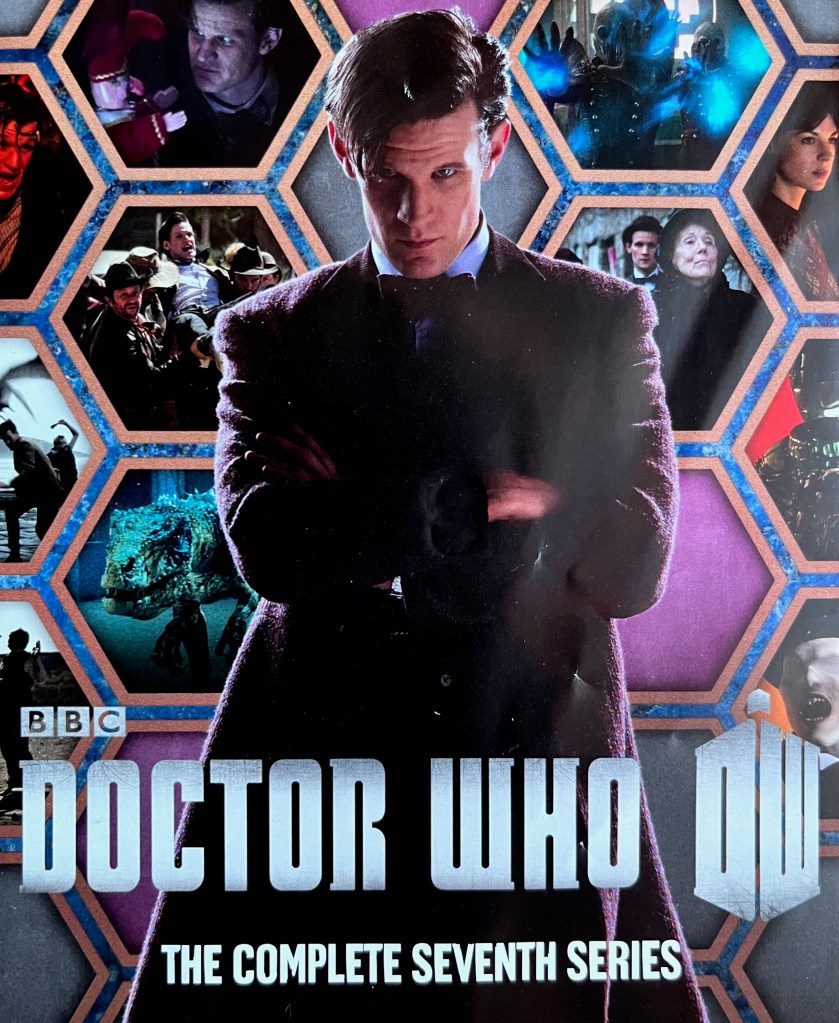 Front cover of the booklet from the old Blu-ray edition of Doctor Who Series 7, showing Matt Smith's Doctor standing with his arms across his chest and a stern expression on his face. Behind him is a collage of large hexagons showing images from the series.