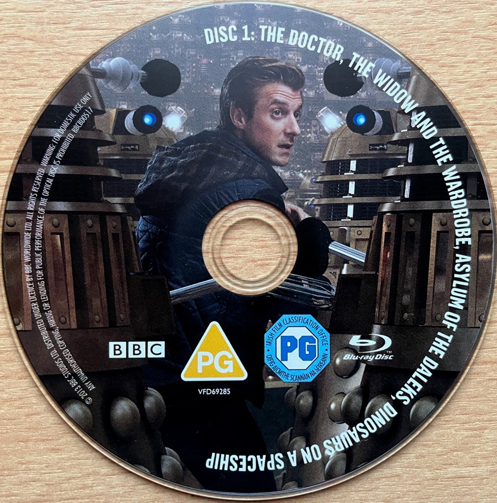 Disc 1 in the Doctor Who Series 7 Blu-ray set, showing an image of Rory surrounded by Daleks. Around the edge of the disc are the episode names The Doctor, The Widow And The Wardrobe, Asylum Of The Daleks and Dinosaurs On A Spaceship, along with copyright information.