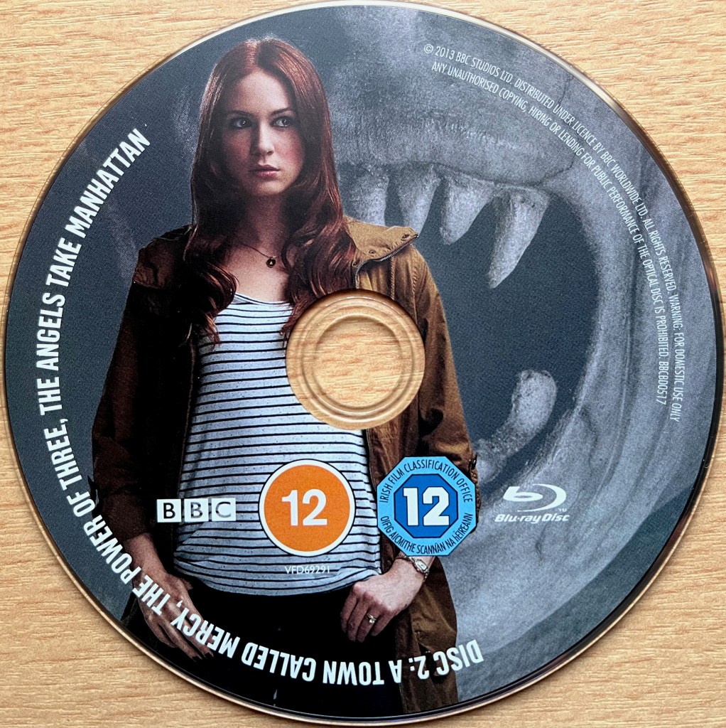 Disc 2 in the Doctor Who Series 7 Blu-ray set, showing an image of Amy in front of a large mouth with pointed teeth. Around the edge of the disc are the episode names A Town Called Mercy, The Power Of Three and The Angels Take Manhattan, along with copyright information.