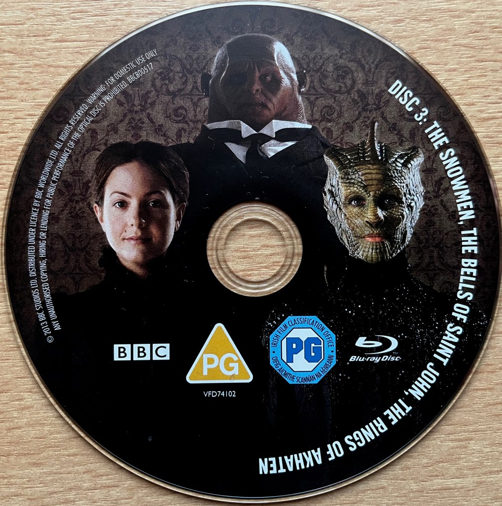 Disc 3 in the Doctor Who Series 7 Blu-ray set, showing an image of Jenny Flint, Strax and Madame Vastra. Around the edge of the disc are the episode names The Snowmen, The Bells Of St John and The Rings Of Akhaten, along with copyright information.