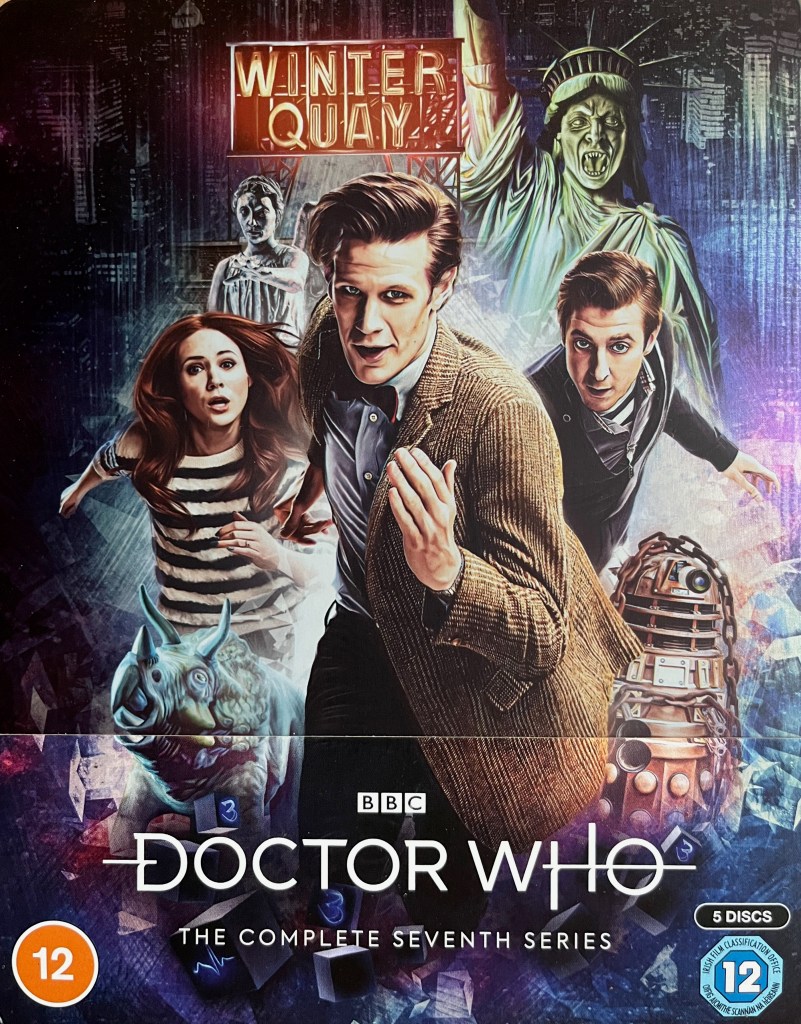 Front cover of the Doctor Who Series 7 Blu-ray steelbook. It shows Matt Smith's Doctor with Amy and Rory running towards us, with a Dalek and 2 Weeping Angels behind them, and a neon sign above them saying Winter Quay.