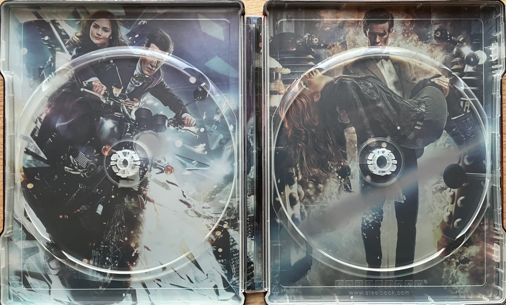 The interior of the Blu-ray steelbook for Doctor Who Series 7, when the discs have been removed. Behind the panels holding the discs are two images. On the left are the Doctor and Clara diving from the sky on a motorbike, while on the right the Doctor is carrying an unconscious Clara in his arms as he walks towards us, with two Daleks in the background.