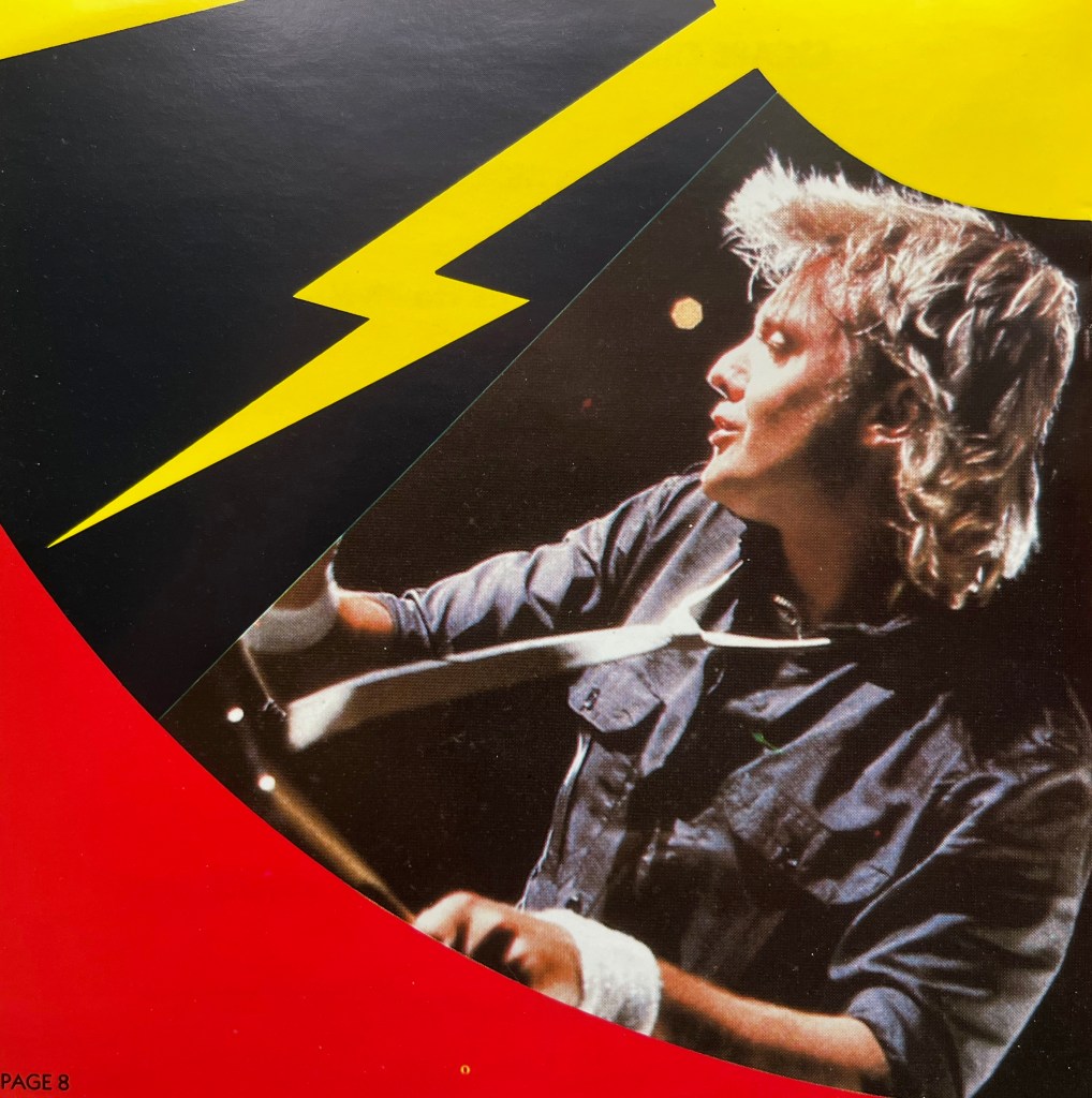 A photo of Roger Taylor playing drums from the booklet with the Flash Gordon album, with a yellow streak of lightning above him.