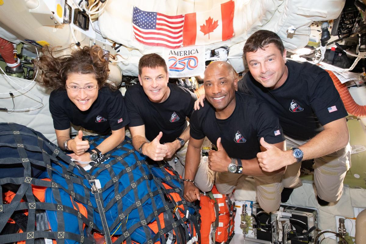The 4 crew members of Artemis 2 floating together inside their capsule, each smiling and giving a thumbs up. From left to right they are Christina Koch, Jeremy Hansen, Victor Glover and Reid Wiseman. Hanging up behind them are an American flag, a Canadian flag, and a white flag with America 250 on it.
