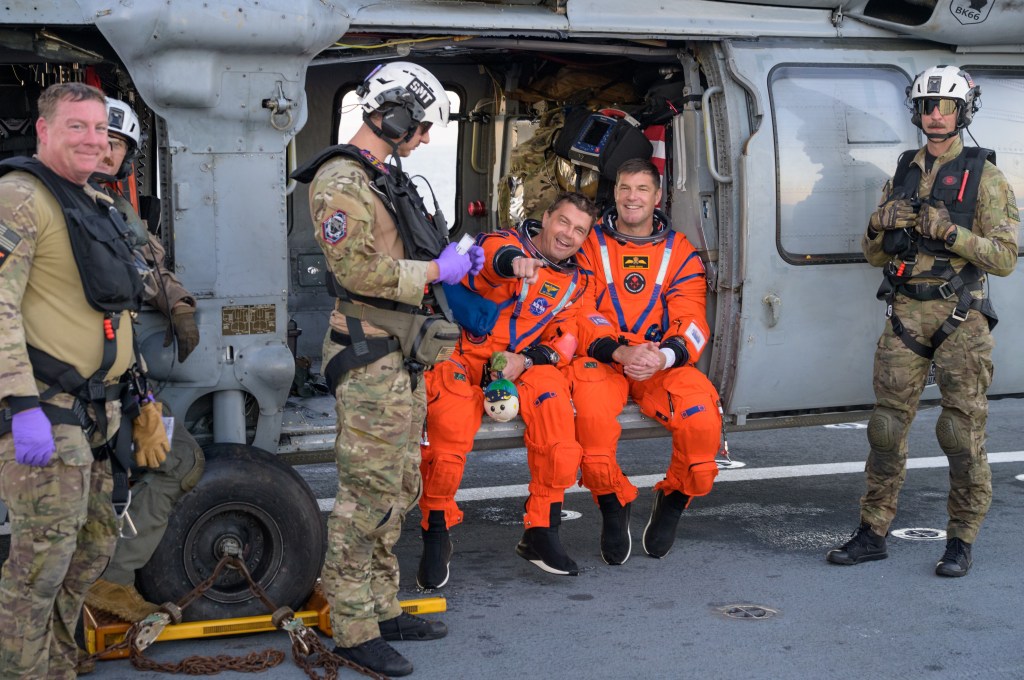 Astronauts Reid Wiseman & Jeremy Hansen wearing orange spacesuits and smiling as they sit in the open doorway of an American Navy helicopter. Reid is pointing towards the camera with one hand, while the other is holding the mascot called Rise, a small white round plush toy with a smiling face. His white face represents the moon, while on his head is a hat representing Earth, with green areas of land surrounded by blue water. The peak of his hat represents the in between area of space with a few yellow stars against a dark background.