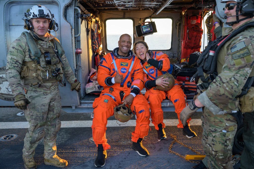 Astronauts Victor Glover and Christina Koch wearing orange spacesuits and smiling as they sit in the open doorway of an American Navy helicopter. Victor is also giving a thumbs up.
