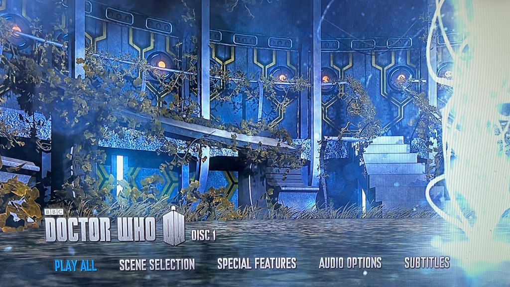 Main menu on the Blu-ray for Doctor Who Series 7. It shows the interior of the Tardis, which has greenery sprouting all over it as if it's been abandoned. On the right is a bright vertical tower of swirling light that stretches from the floor to the ceiling. At the bottom of the screen is the Doctor Who logo, along with options for Play All, Scene Selection, Special Features, Audio Options and Subtitles.