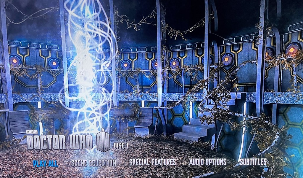 Main menu on the Blu-ray for Doctor Who Series 7. It shows the interior of the Tardis, which has greenery sprouting all over it as if it's been abandoned. On the left is a bright vertical tower of swirling light that stretches from the floor to the ceiling. At the bottom of the screen is the Doctor Who logo, along with options for Play All, Scene Selection, Special Features, Audio Options and Subtitles.