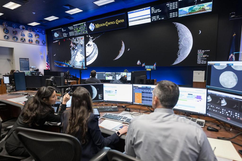 A view inside Mission Control Center at NASA, where operators are sitting at computers and looking up at big screens showing views of the moon from the Orion spaceship, with Earth looking very small in the background by comparison.