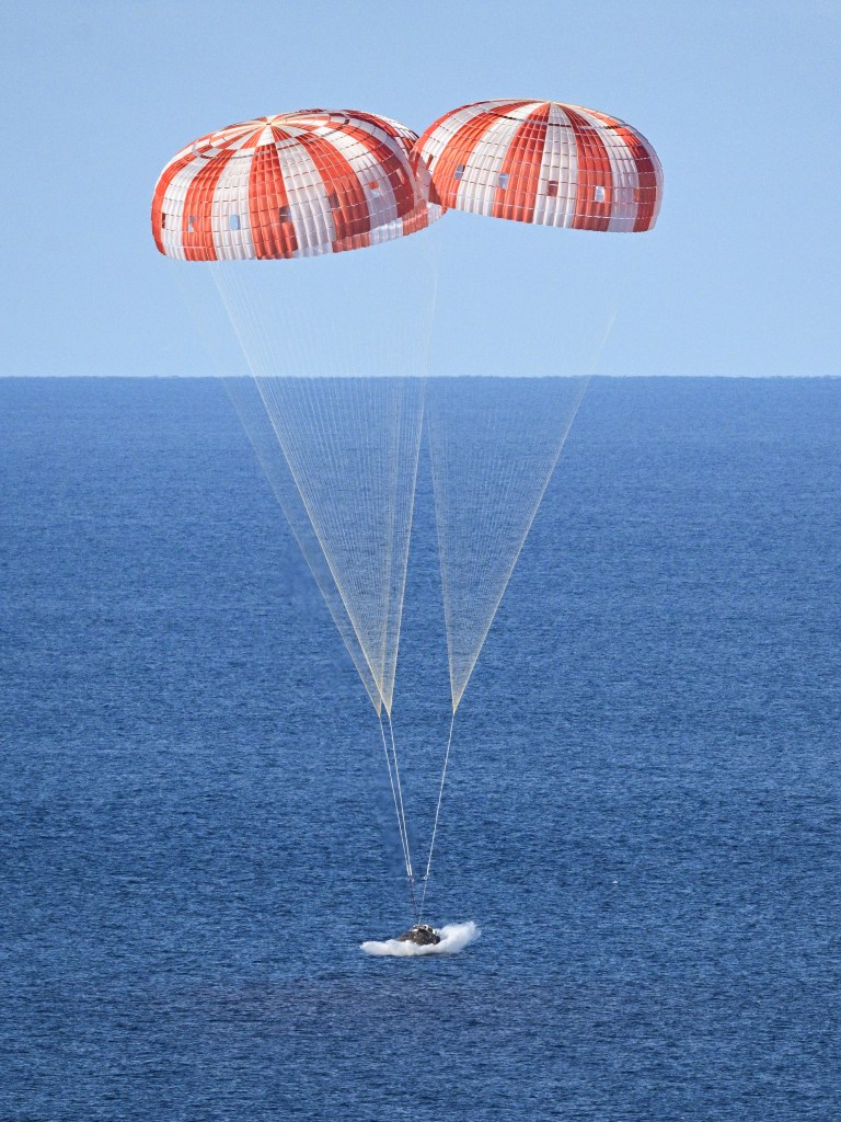 The Orion space capsule splashing down in the Pacific Ocean, supported by 3 large orange and white striped parachutes.