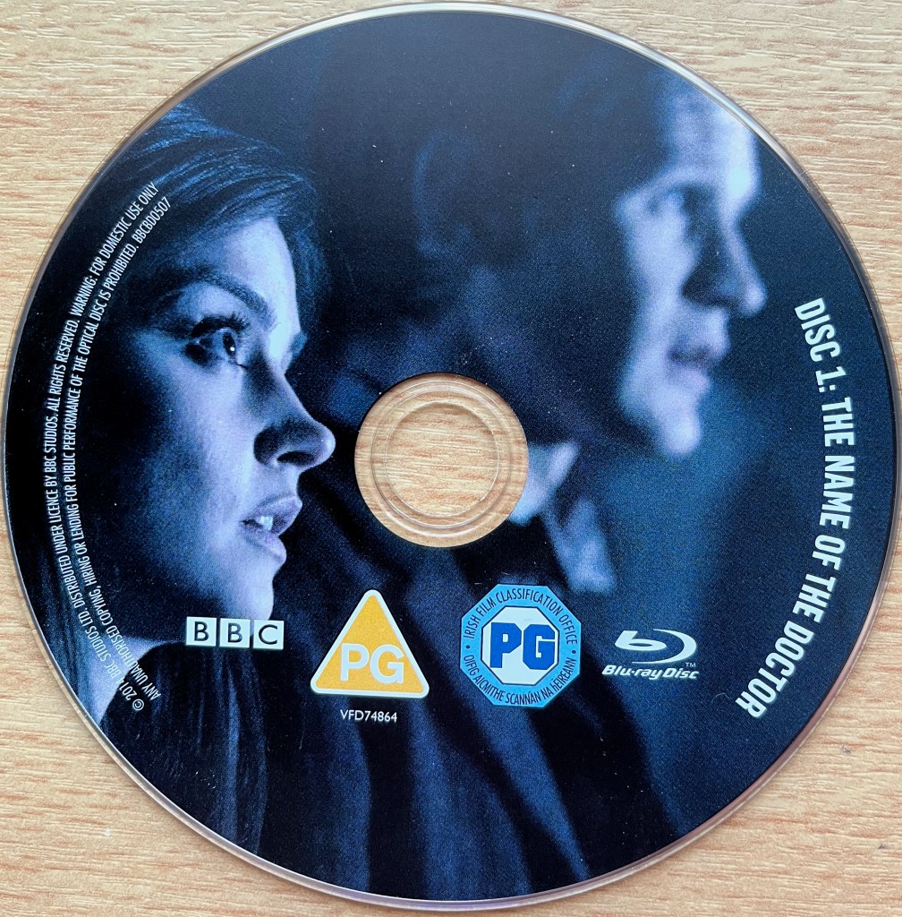 Disc 1 in the Doctor Who 50th Anniversary Specials Blu-ray set, showing an image of Clara and Matt Smith's Doctor, looking to the right with concerned expressions. Around the right edge of the disc is the episode title The Name Of The Doctor, along with copyright information on the left edge.