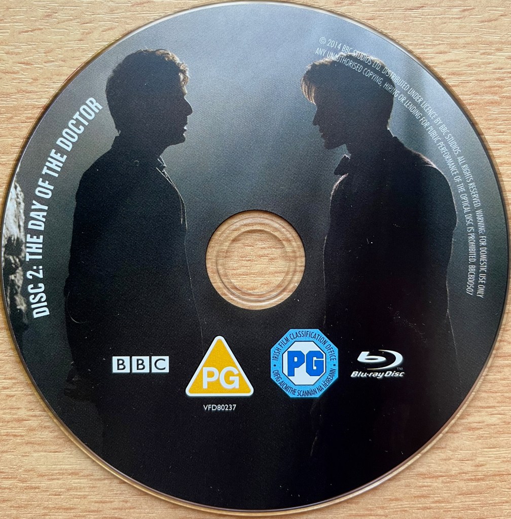 Disc 2 in the Doctor Who 50th Anniversary Specials Blu-ray set, showing an image of David Tennant and Matt Smith's Doctors facing each other in a shadowy space that makes them almost look like silhouettes. Around the left edge of the disc is the episode title The Day Of The Doctor, along with copyright information on the right edge.