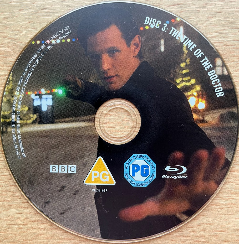 Disc 3 in the Doctor Who 50th Anniversary Specials Blu-ray set, showing an image of Matt Smith's Doctor facing us and pointing his sonic screwdriver at us with a green light on the front of it. Behind him is an out of focus background showing the Tardis on the left and a lit-up Christmas tree on the right. Around the right edge of the disc is the episode title The Time Of The Doctor, along with copyright information on the left edge.