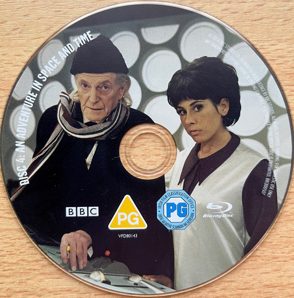 Disc 4 in the Doctor Who 50th Anniversary Specials Blu-ray set, showing an image of David Bradley portraying the first Doctor, with Susan by his side, in the old style Tardis with white circles making up the wall behind them. Around the left edge of the disc is the episode title An Adventure In Space And Time, along with copyright information on the right edge.