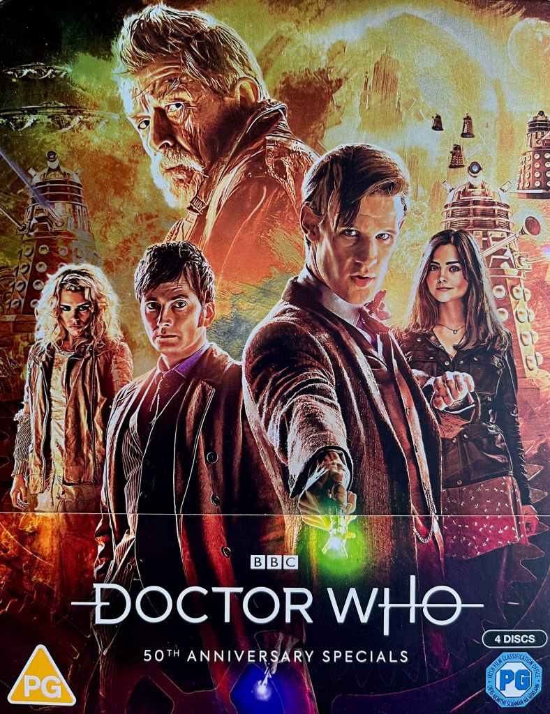 Front cover of the Doctor Who 50th Anniversary Specials Blu-ray steelbook. It shows Matt Smith, David Tennant and John Hurt as their respective iterations of the Doctor, along with companions Clara Oswald and Rose Tyler, while Daleks and their spaceships swarm around in an explosive, fiery background, in amongst which is the large dome on the planet Gallifrey.