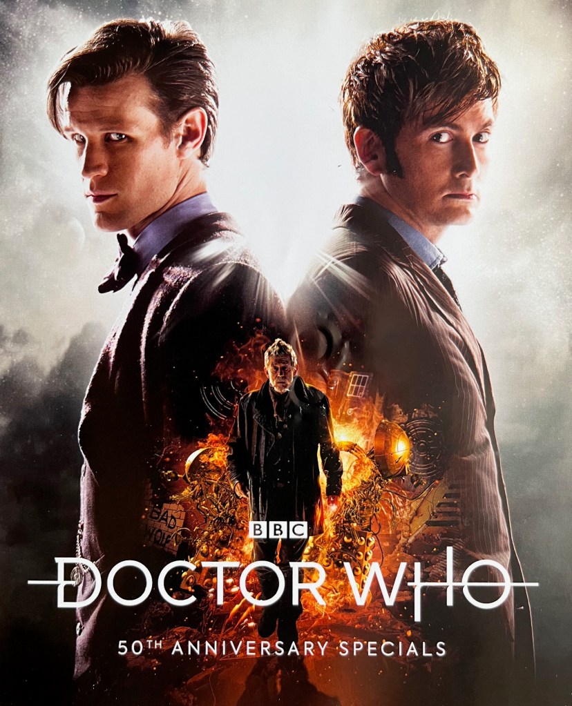 Front side of the leaflet included with the Doctor Who 50th Anniversary Specials Blu-ray Steelbook, featuring a photo of Matt Smith's and David Tennant's Doctors standing back to back, turning their heads to face towards us.. In the middle, in a smaller image lower down the page, John Hurt's Doctor strides away from an explosion, in which the Daleks and the Tardis are visible.