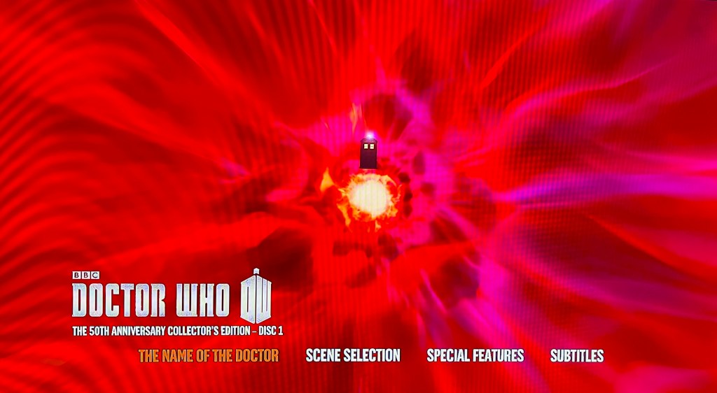 The Blu-ray menu for the Doctor Who 50th Anniversary Specials, showing the Tardis flying through a swirling tunnel of red, pink and yellow colours that form the time vortex. Across the bottom of the screen is the Doctor Who logo, the disc title of the 50th Anniversary Collector's Edition Disc 1, and the options for The Name Of The Doctor, Scene Selection, Special Features and Subtitles.