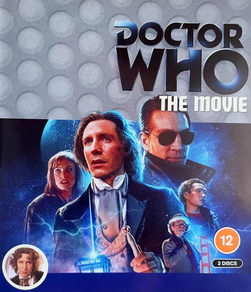 Alternate front cover for the Blu-ray of Doctor Who The Movie, designed to match previous Classic era releases. The top half of the cover has the Doctor Who logo from the movie over a grey background with lighter grey circles embedded into it, while the bottom half shows Paul McGann as the 8th Doctor, along with his companion Grace, the Master and his companion Chang Lee, and Sylvester McCoy as the previous incarnation of the Doctor, while a planet floats in space behind them. A second image of Paul McGann's face is shown in a circle in the very bottom left corner.