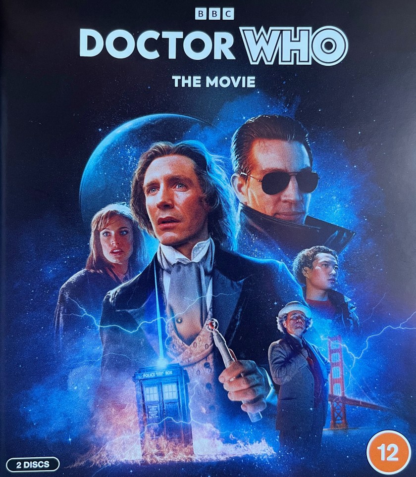 Front cover for the Blu-ray of Doctor Who The Movie, showing Paul McGann as the 8th Doctor, along with his companion Grace, the Master and his companion Chang Lee, and Sylvester McCoy as the previous incarnation of the Doctor. In the background is a planet floating in space, and at the bottom is a small image of the Tardis surrounded by lightning.