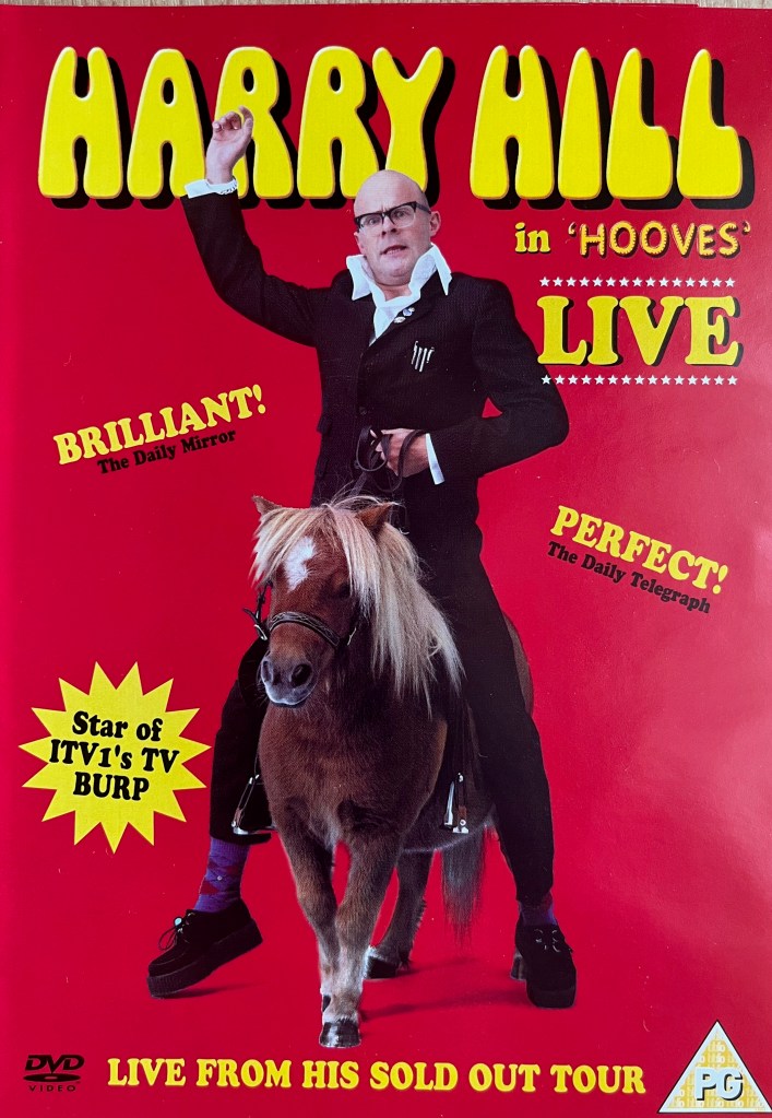 Front cover of Harry Hill's Hooves DVD. Against a red background, Harry is riding a small horse, his right arm up in the air while his left hand holds the reins. At the top of the cover is his name in large yellow letters, while smaller text to the right of him says In Hooves Live.