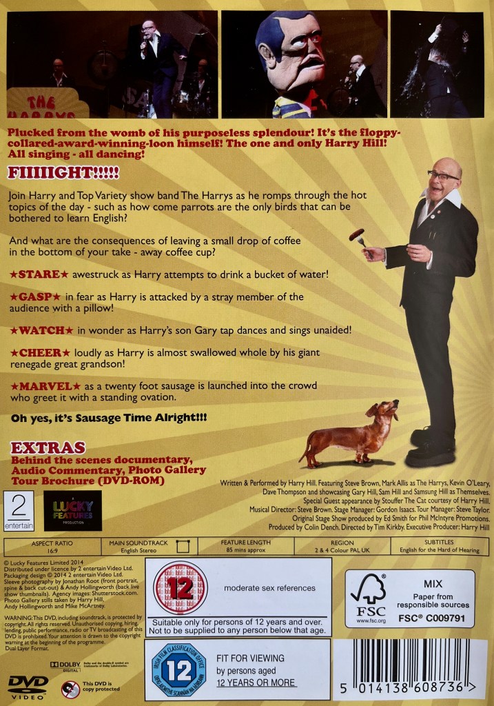 Back cover of the DVD for Harry Hill's Sausage Time. At the top, above the description of the DVD, are a few images of Harry on stage during the show, while to the right of the description is an image of Harry standing while holding a sausage on a fork, as a small dog looks up longingly.