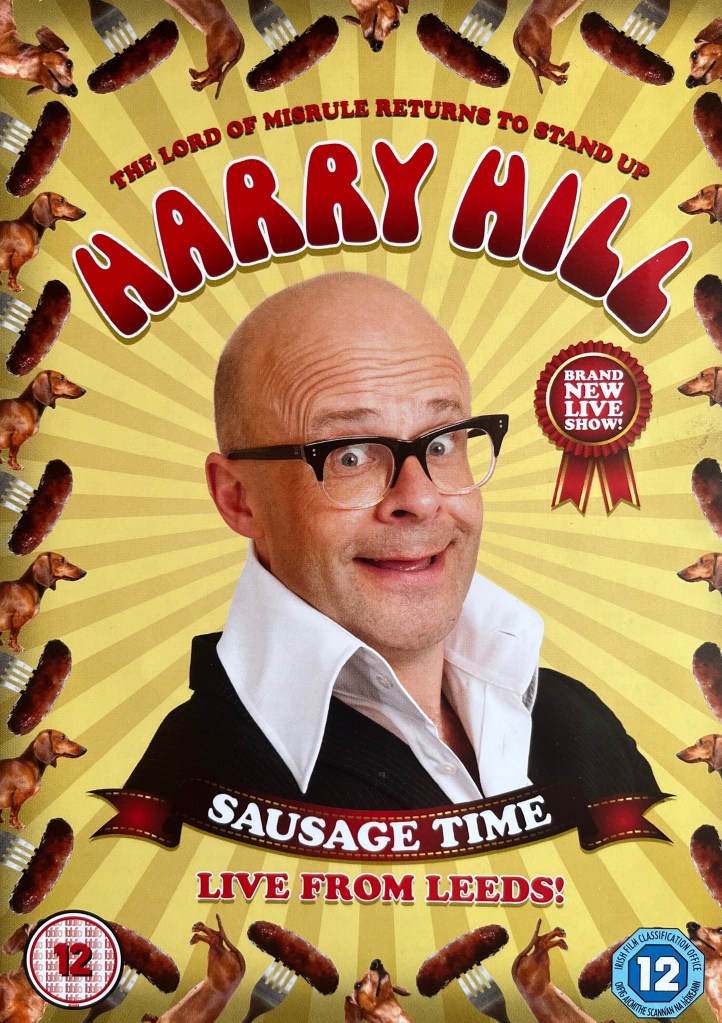 Front cover of the DVD for Harry Hill's Sausage Time. In the centre is a head and shoulders image of Harry smiling, against a background of light yellow and orange stripes that emanate from around his head like rays of light. His name is above that, in red letters with a white outline, and above that is smaller text that reads The Lord of Misrule returns to stand up. Below his image is the text Sausage Time, live from Leeds. The border all around the cover consists of alternating images of sausages on forks and the head or tail ends of dogs.