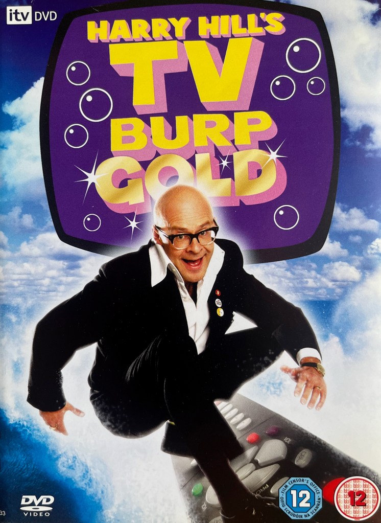 Front cover of the DVD for Harry Hill's TV Burp Gold. The title is shown in large yellow letters on a TV screen with curved edges, which has a dark purple background and white bubbles floating inside it. Below the screen, Harry is surfing on a large remote control.