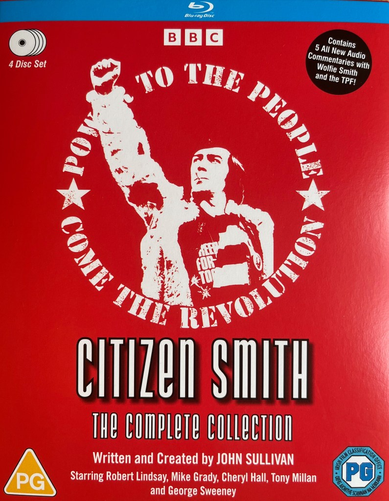 Front cover for the Blu-ray box set of Citizen Smith, which has a red background. It shows a white filtered image of Wolfie wearing his Freedom for Tooting t-shirt as he pumps his first triumphantly into the air. Surrounding him in a circle is white text that says Power To The People, Come The Revolution. Below that is text that says Citizen Smith, The Complete Collection. Written and created by John Sullivan. Starring Robert Lindsay, Mike Grady, Cheryl Hall, Tony Millan and George Sweeney. At the top right of the cover is a black circle with white text that says the set contains
5 All New Audio
Commentaries with
Wolfie Smith and the TPF.