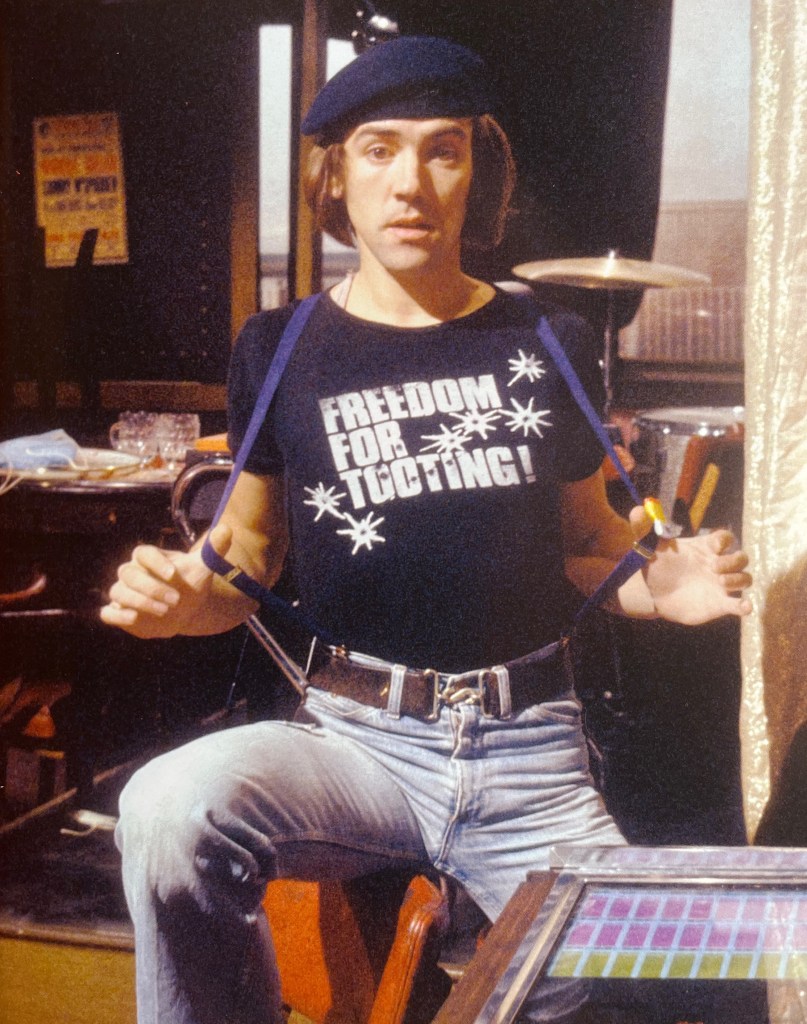 A photo of Wolfie Smith from Citizen Smith. Sitting on a chair, he's wearing a black t-shirt with the words Freedom For Tooting in large white capital letters surrounded by white splodges. He's using his thumbs to pull forward each side of his braces, which are attached to his light blue denim jeans, and on his head he's wearing a dark blue beret.