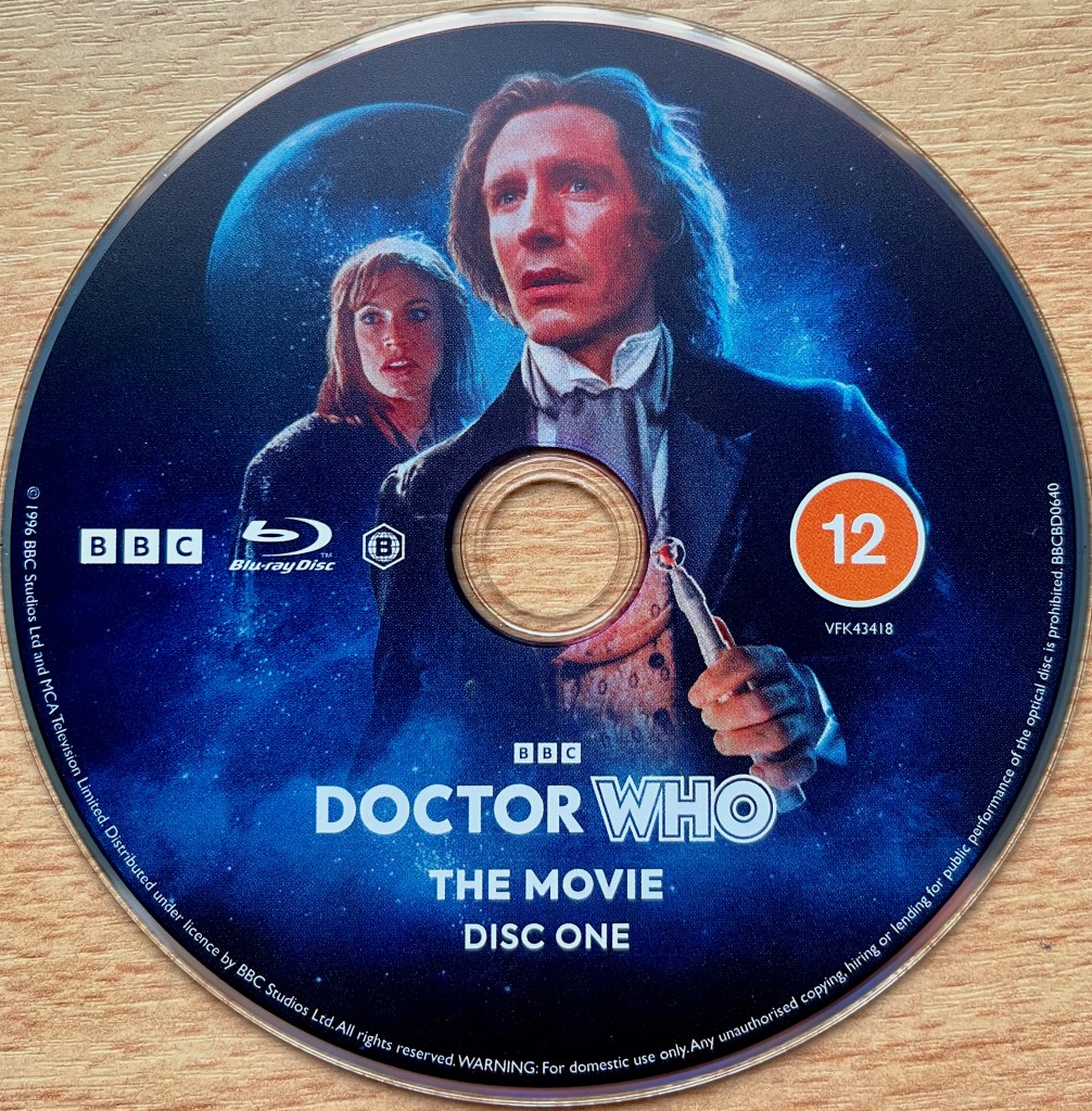 Blu-ray disc 1 for Doctor Who the movie, showing Paul McGann as the 8th Doctor with companion Grace, while a planet floats in space in the background.