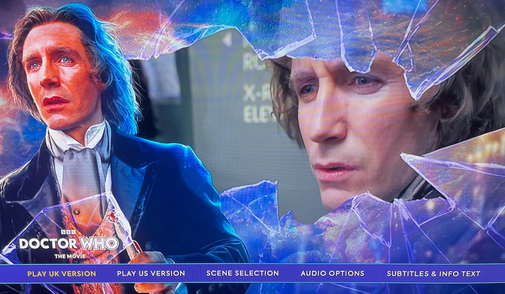 Blu-ray menu from the first disc for Doctor Who The Movie. It shows a photo of Paul McGann as the 8th Doctor on the left, while the rest of the screen shows a large area of smashed glass, through which we also see the face of the Doctor. Across the bottom of the screen are menu options to play the UK and US versions of the film, along with Scene Selection, Audio Options and Subtitles.