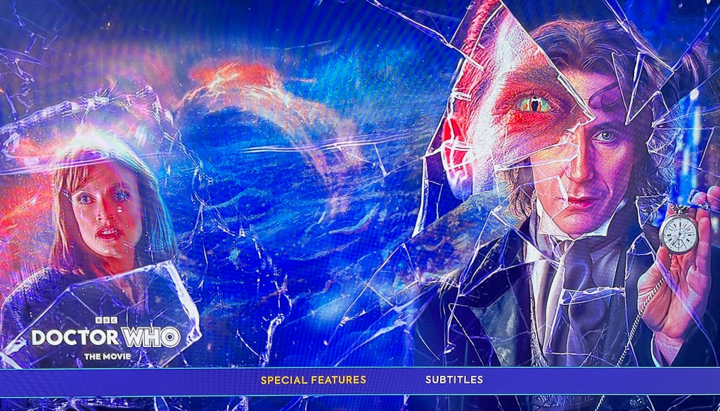 Blu-ray menu for Disc 2 of the Doctor Who Movie. On the right, looking through a smashed area of the screen, is Paul McGann as the 8th Doctor holding up a pocket watch. On the left, against a background of the universe, is an image of his companion Grace. At the bottom of the screen is a menu bar with options for Special Features and Subtitles.