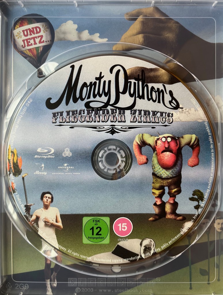 Blu-ray disc for Monty Python’s Fliegender Zirkus. The scene below the title shows a man holding a tall pole with a flame on the top on the left, while on the right is an angry looking man with a very red face, and his face is so big that his chin covers the top half of his chest. At the top of the disc, the tip of a thumb and forefinger look like they are about to pinch the title of the show, and when placed in the case they line up with the full artwork of the hand in the background of the steelbook.