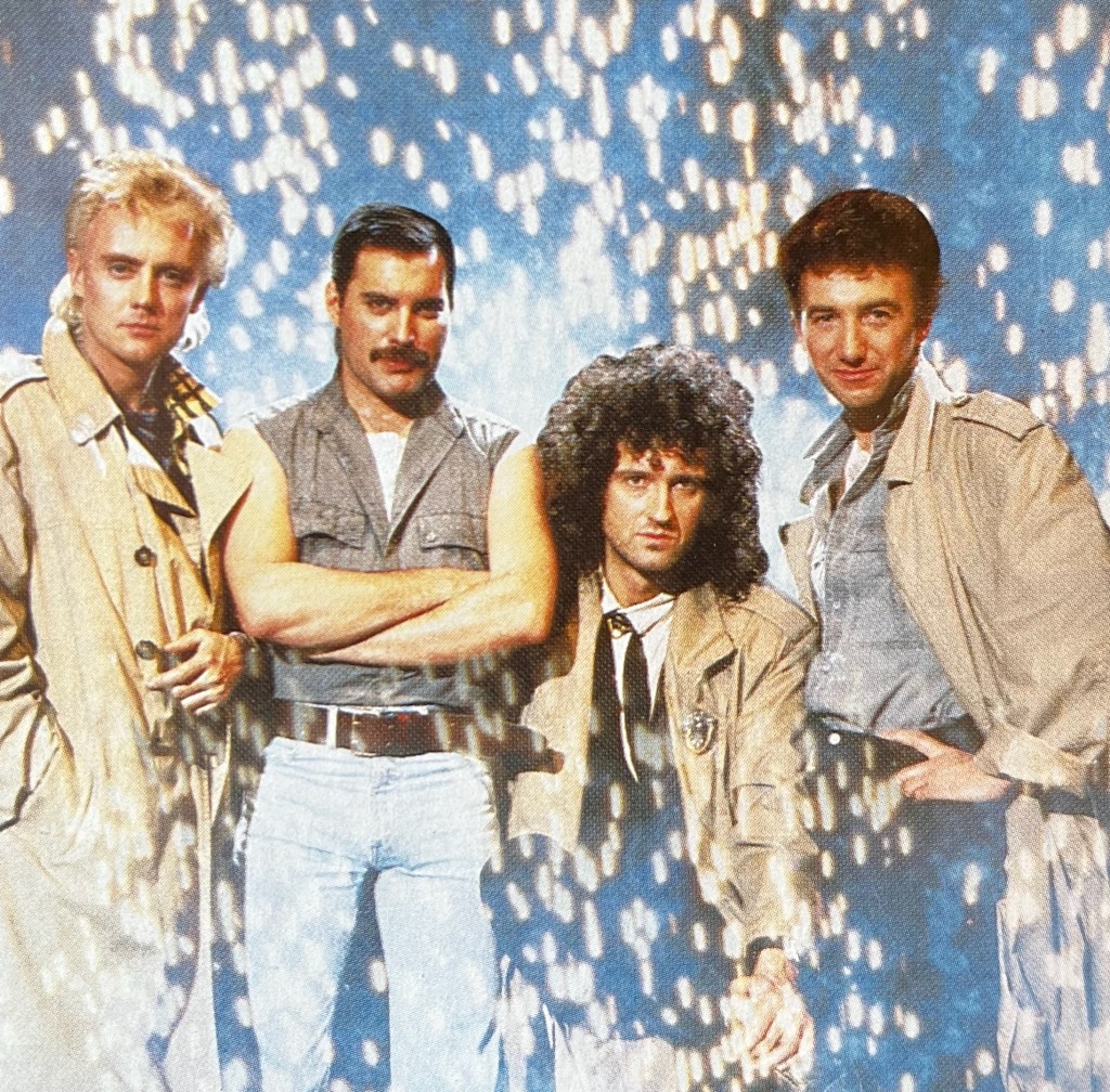 A photo of Queen members Roger, Freddie, Brian and John standing together, while lots of bright yellow sparks rain down behind them.