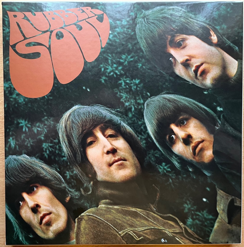 Front cover of the box set for the Beatles album Rubber Soul, showing the 4 members of the band against a green leafy background. The arrangement of their faces moves from bottom left to top right in a curved formation. The album title takes up the top left corner, with the word Rubber above the word Soul. The letters are bulging, as if like rubber being blown up, with the word Soul being the largest, the bottom part of each letter bulging out significantly.